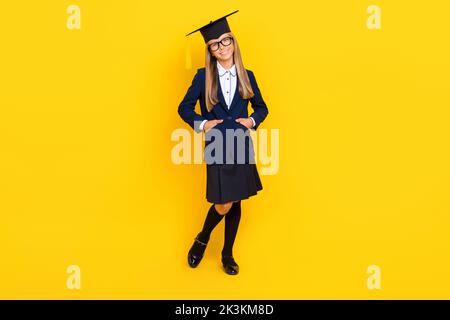 Photo pleine grandeur de jolie jolie petite fille mignonne avec une coiffure droite habillé bleu uniforme bras dans les poches isolées sur fond jaune Banque D'Images