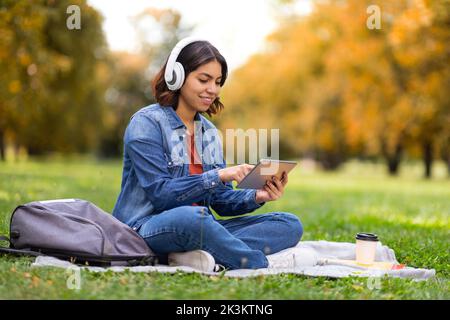 Détente en plein air. Jeune femme arabe souriante portant un casque sans fil à l'aide d'une tablette numérique Banque D'Images