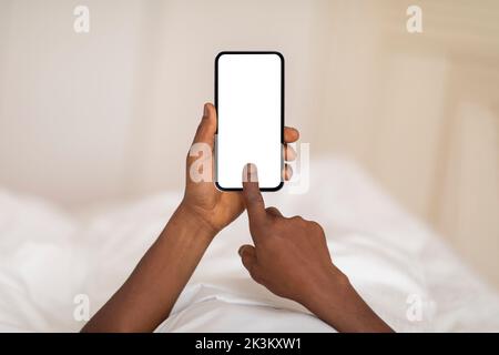 Homme noir touchant l'écran vide du smartphone en étant allongé dans le lit à la maison Banque D'Images