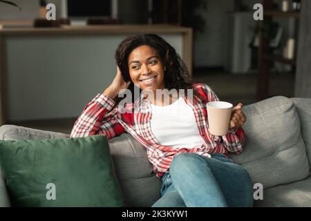 Bonne jeune femme afro-américaine avec une tasse de boisson préférée, aime la paix, le silence et le confort Banque D'Images