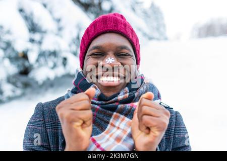 homme de main afro-américain en chapeau rouge regarder la caméra avec la neige tasseuse, sourire blanc et flocon de neige sur le nez à l'extérieur dans le parc Banque D'Images