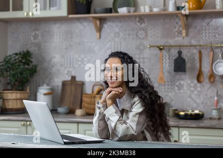 Portrait d'une jeune belle femme hispanique à la maison, femme indépendante travaillant à distance à l'aide d'un ordinateur portable, regardant l'appareil photo et souriant dans des lunettes et des cheveux bouclés dans la cuisine Banque D'Images