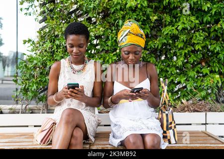 Adultes Afro-américaines amies dans des robes élégantes et des accessoires authentiques assis sur le banc dans le parc de la ville et en utilisant des smartphones Banque D'Images