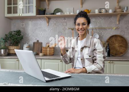 Portrait d'une jeune femme hispanique à la maison, femme d'affaires souriant et regardant l'appareil-photo agitant geste de salutation, indépendant dans la cuisine utilisant un ordinateur portable pour le travail à distance. Banque D'Images