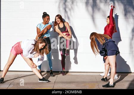 Groupe de jeunes amies actives multiethniques gaies dans les vêtements de sport souriant et se réchauffant près du mur blanc dans la rue pendant l'entraînement en plein air au soleil Banque D'Images