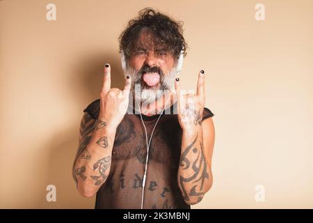 Rocker rebelle et mature avec tatouages écoutant de la musique dans les écouteurs et regardant la caméra tout en montrant la langue et le geste de corne sur le dos beige Banque D'Images