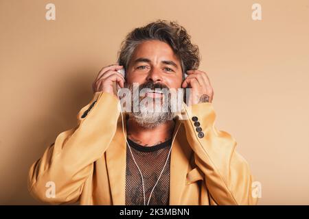Contenu Homme d'âge moyen avec barbe grise écouter des chansons dans l'oreillette tout en regardant l'appareil photo sur fond beige en studio Banque D'Images