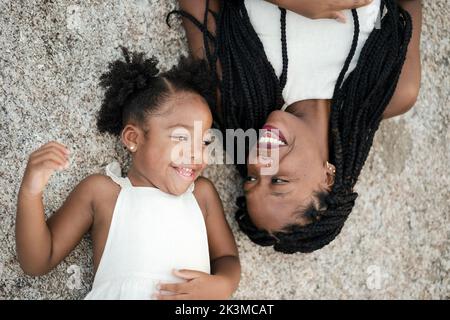 Vue de dessus de bonne maman noire et fille dans des vêtements blancs souriant et regardant l'un l'autre tout en étant allongé sur la surface de pierre ensemble Banque D'Images
