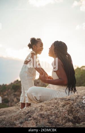 Bonne maman afro-américaine avec des cheveux bouclés tenant les mains de la fille et parlant tout en étant assis sur le rocher contre le ciel nuageux de soleil en fin de semaine dans la nature Banque D'Images