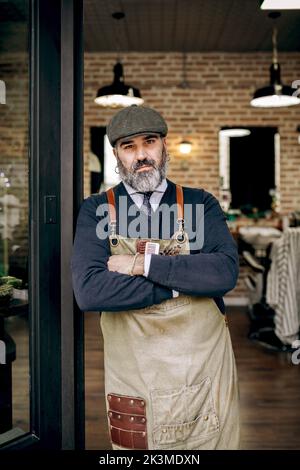 Portrait d'un coiffeur homme mûr et confiant en tablier avec une barbe debout avec des bras croisés à l'entrée du salon de coiffure et regardant l'appareil photo Banque D'Images