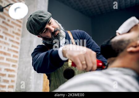 Utilisez un rasoir électrique pour couper une barbe noire d'un client ethnique masculin dans un salon de coiffure traditionnel Banque D'Images