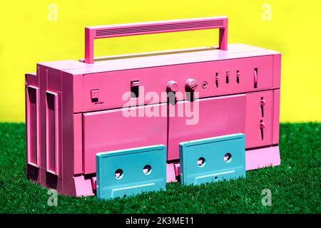 Lecteur de cassettes vintage rose vif avec deux cassettes bleues placées sur de l'herbe verte sur fond jaune en studio Banque D'Images