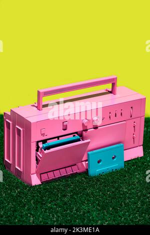 Lecteur de cassettes vintage rose vif avec cassettes bleues placées sur de l'herbe verte sur fond jaune en studio Banque D'Images