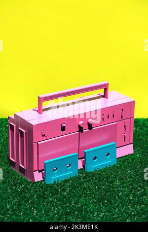 Lecteur de cassettes vintage rose vif avec deux cassettes bleues placées sur de l'herbe verte sur fond jaune en studio Banque D'Images