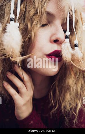 Sereine blonde femme en robe rouge et avec les yeux fermés montrant le chat de rêve décoratif avec des plumes sur fond rose en studio Banque D'Images