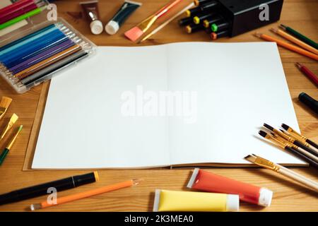 Maquette d'artiste créatif sur table en bois . Pose à plat, vue de dessus avec carnet d'esquisses vierge avec espace de copie, crayons, pinceaux et peintures. Art Banque D'Images