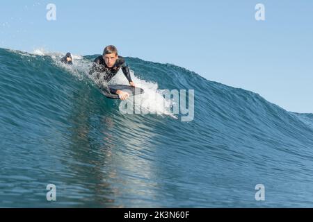 Un jeune homme en combinaison noire attrape une vague sur un bodyboard de planche de boogie à Piha Beach, Auckland, Nouvelle-Zélande. Banque D'Images