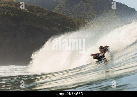 Un jeune homme en combinaison noire attrape une vague sur un bodyboard de planche de boogie à Piha Beach, Auckland, Nouvelle-Zélande. Banque D'Images