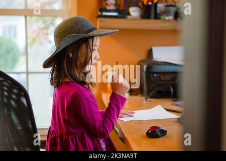 Un petit enfant en chapeau est assis au bureau avec du papier et un crayon Banque D'Images