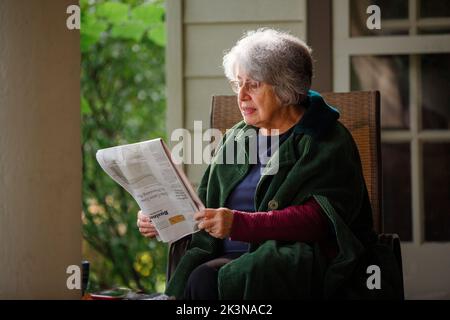 Une femme âgée est assise sur le porche avant dans un journal de lecture en manteau Banque D'Images