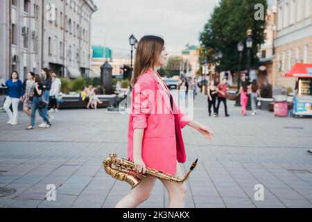 une fille dans une veste rose avec un saxophone marche le long de la rue de la ville Banque D'Images