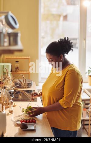 Portrait vertical de la femme noire en surpoids cuisant un repas sain dans la cuisine éclairée par la lumière du soleil Banque D'Images