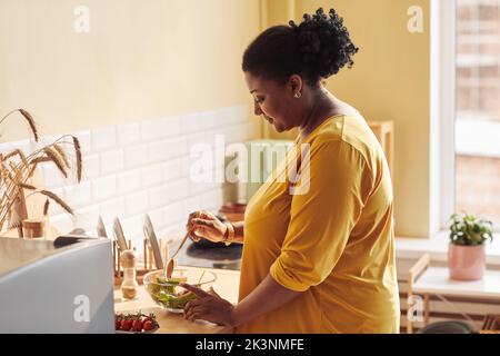 Vue latérale portrait d'une femme noire en surpoids cuisant un repas sain dans la cuisine éclairée par la lumière du soleil, espace de copie Banque D'Images