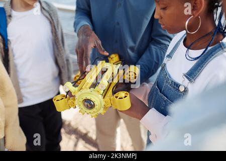 Gros plan de la jeune fille noire tenant le modèle de robot pendant la classe d'ingénierie à l'extérieur avec l'aide d'enseignant Banque D'Images