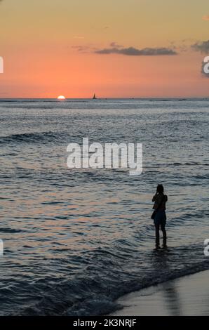 Une femme seule sur la plage de Waikiki à Hawaï prenant une photo du soleil couchant sur l'océan Pacifique Banque D'Images