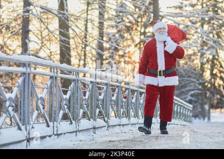 Portrait complet du sac de transport traditionnel du Père Noël avec cadeaux et marche vers l'appareil photo en hiver, espace copie Banque D'Images
