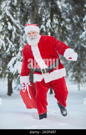 Portrait vertical sur toute la longueur du sac de transport traditionnel du Père Noël avec des cadeaux dans la forêt d'hiver et courant vers l'appareil photo Banque D'Images
