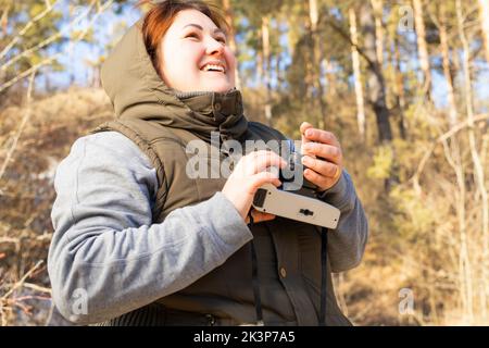 Une femme joyeuse en plein air vêtue de vêtements chauds d'hiver qui garde l'appareil photo entre les mains et qui profite de la nature le jour d'hiver ensoleillé. Banque D'Images