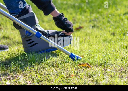 Un garçon avec un pied cassé et une chaussure orthopédique ou un marcheur après une fracture osseuse repose dans un parc public sur un banc en herbe verte pour recréer et réhabiliter Banque D'Images