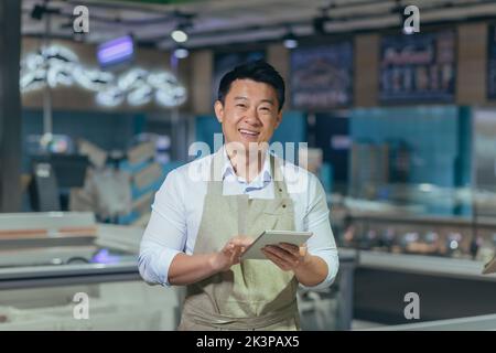 Portrait d'un beau jeune homme asiatique, employé d'un magasin, d'un supermarché. Debout dans un tablier, tenant un bloc-notes, regardant la caméra, souriant Banque D'Images