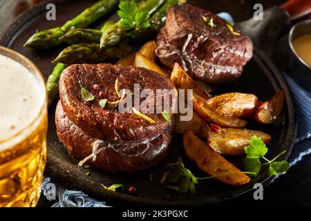 De délicieux médaillons de bœuf avec des pommes de terre frites et des asperges cuites à la vapeur servis pour le dîner sur une poêle à frire près d'une tasse de bière froide Banque D'Images