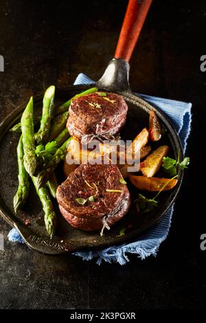 Des médaillons de viande avec asperges et pommes de terre pour le dîner servis sur une poêle rustique sur une serviette bleue sur fond noir Banque D'Images