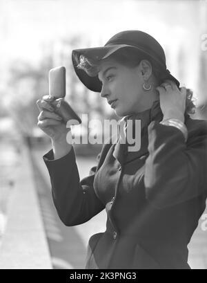 Maquillage dans le 1950s. Une jeune femme tient un miroir de poche et fixe un détail dans ses cheveux. Le modèle tendance porte une veste avec une ceinture fine visible, un chapeau assorti et une écharpe autour de son cou. Suède 1952 Kristoffersson Réf. 38K-7 Banque D'Images