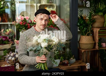 Bon homme floriste de style caucasien portant un chandail, un bonnet et des lunettes tenant un bouquet de fleurs. Concept de petite entreprise. Mâle barbu avec bouquet de roses blanches dans un magasin de fleurs. Image de haute qualité Banque D'Images