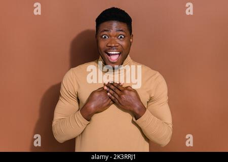 Photo du jeune brunet impressionné gars paumes poitrine porter beige turtleneck isolé sur fond brun Banque D'Images