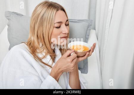 Jolie femme caucasienne en peignoir blanc buvant du thé au citron chaud en profitant des procédures de spa dans le centre de bien-être Banque D'Images