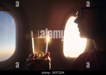 Homme prenant un verre pendant le vol. Passager tenant un verre de vin mousseux contre la fenêtre de l'avion au coucher du soleil. Banque D'Images