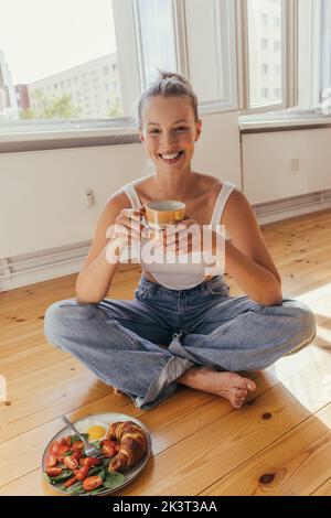 Femme blonde souriante en Jean et haut tenant la tasse près de savoureux petit déjeuner sur le sol à la maison, image de stock Banque D'Images