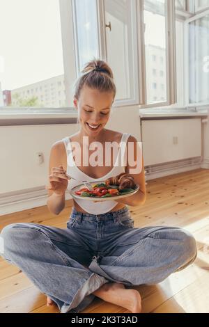 Femme positive en haut et Jean tenant une assiette avec un délicieux petit déjeuner sur le sol à la maison, image de stock Banque D'Images