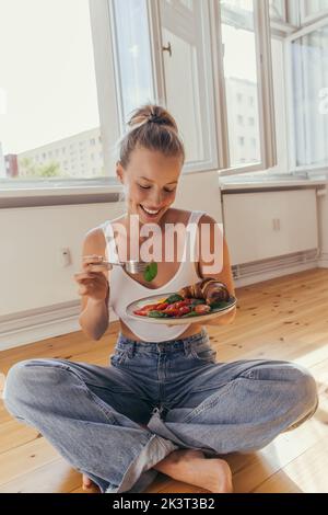 Jeune femme souriante tenant un délicieux petit déjeuner sur l'assiette tout en étant assise sur le sol à la maison, image de stock Banque D'Images