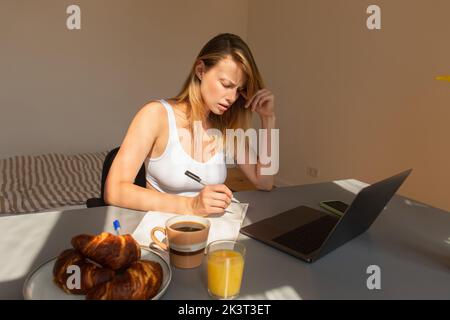 Inquiet freelance écrivant sur ordinateur portable près des appareils et petit déjeuner sur table à la maison, image de stock Banque D'Images