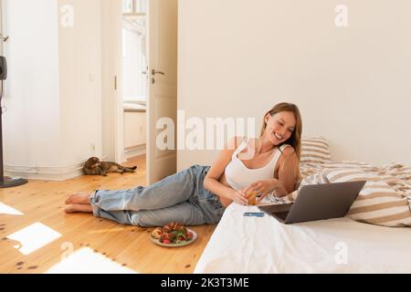 Jeune femme tenant du jus d'orange près de l'ordinateur portable et le petit déjeuner dans la chambre, image de stock Banque D'Images