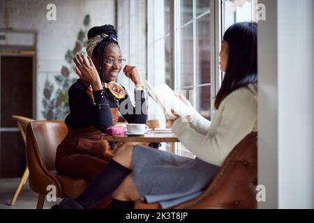Deux filles d'université multiculturelles heureuses sont assises à la cafétéria et étudient pour un examen. Banque D'Images