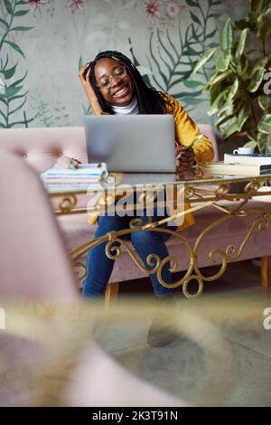 Une jeune fille d'université africaine souriante s'assoit dans un café et termine le projet scolaire sur son ordinateur portable. Banque D'Images
