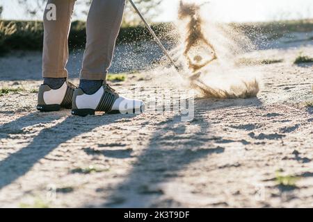 Vue latérale au niveau du sol de la récolte anonyme athlète masculin dans les chaussures de golf frapper balle avec putter dans la zone de sable dangereux du terrain Banque D'Images