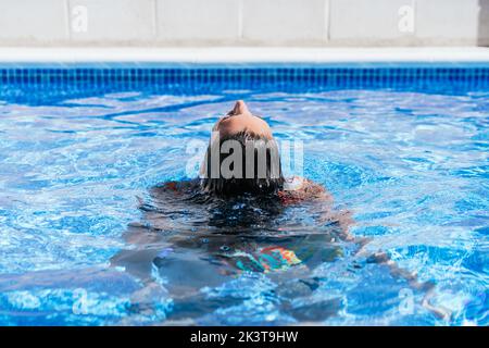 Vue arrière de la jeune femme détendue dans le bikini coloré flottant avec les yeux fermés dans l'eau de cristal de la piscine bleue tout en appréciant les vacances d'été Banque D'Images